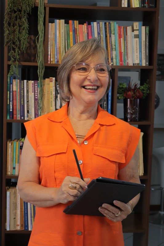 Professora Rosane Reis sorrindo, segurando um tablet, com estante de livros ao fundo